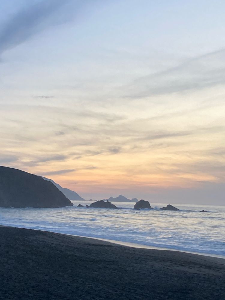 pale white, gray & pink clouds in a hazy sky at sunset over a slightly choppy, frothy white ocean, waves breaking at the shore rolling in past two rows of rock formations