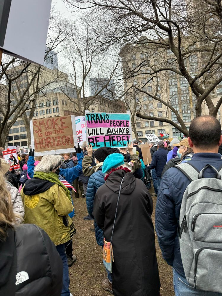 A crowd holding signs with a pink white and blue sign in the middle that says “trans people will always be here”
