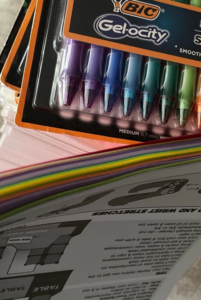 A box of rainbow gel pens and a stack of printed paper that is also multi-colored
