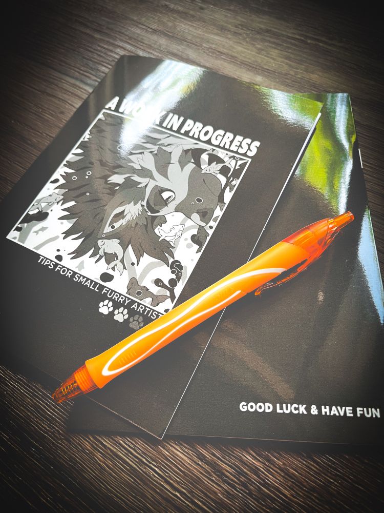 A booklet and orange pen. 

The front of the booklet says: A Work In Progress, Tops for small furry artists
The back of the booklet says: Good Luck & Have afun