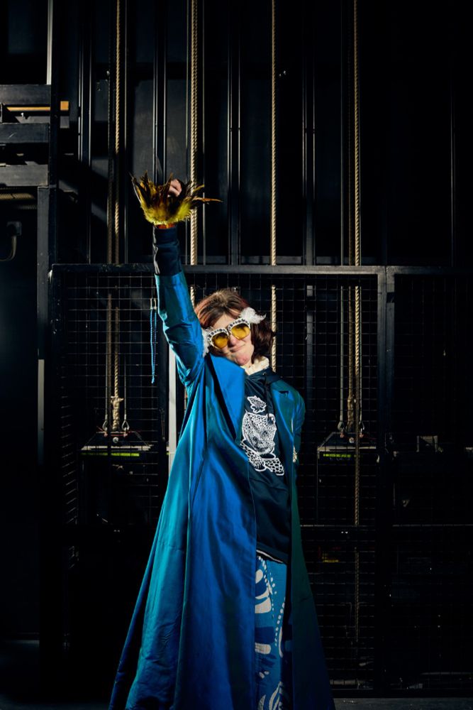 Lung Ha actor Liza Gogoliuk poses for the camera with her right hand stretched straight up in the air in a defiant pose. Liza is wearing a full-length dark blue coat; a navy top with a black-and-white bear design on the front; light blue trousers with a white and darker blue pattern across it; and tinted sunglasses with amber lenses, a white frame and tufts of white feathers where the frame meets the glasses’ arms/temples. A larger tuft of yellow and black feathers can also be seen emerging from the sleeve of her outstretched right hand. Behind Liza we can see a black wall and pulley ropes backstage in a theatre space.