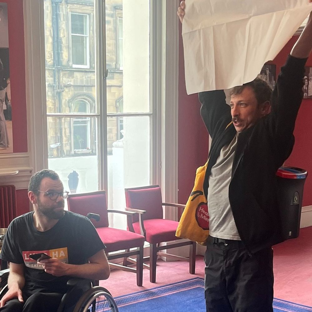 I'm Me Lead Artist Gavin and Lead Facilitator Ryan in rehearsal for their performance at the I'm Me Festival. Ryan is holding a makeshift placard above his head while Gavin looks over to him.