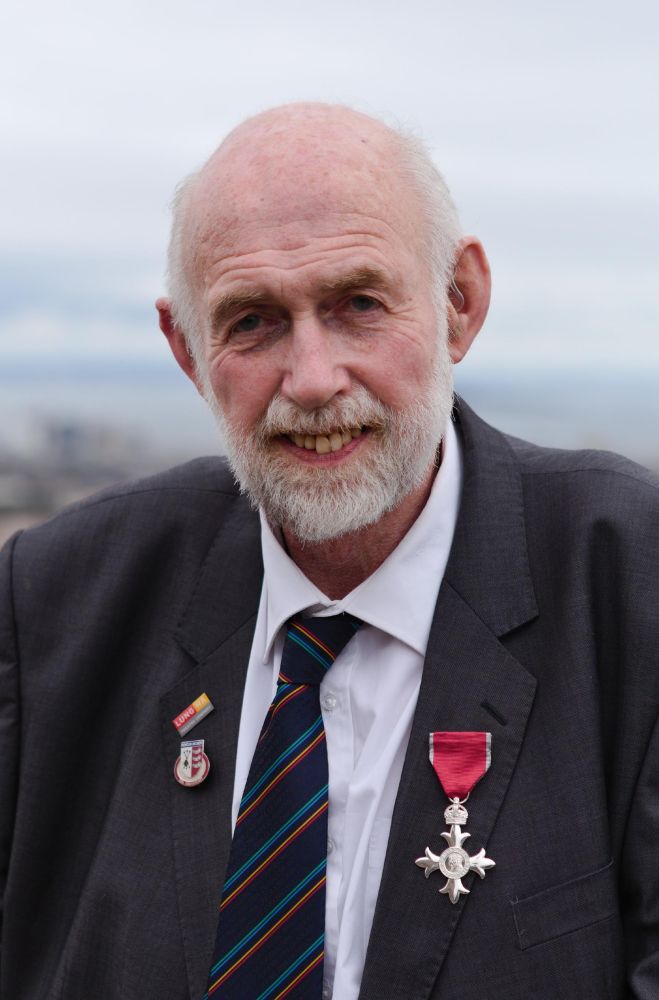 Company co-founder and Honorary President Richard Vallis poses for the camera with his recently-awarded MBE medal pinned to the left lapel of his jacket. The right lapel boasts 2 additional pins depicting the Lung Ha Theatre Company logo as well as the Penicuik Archery coat of arms.