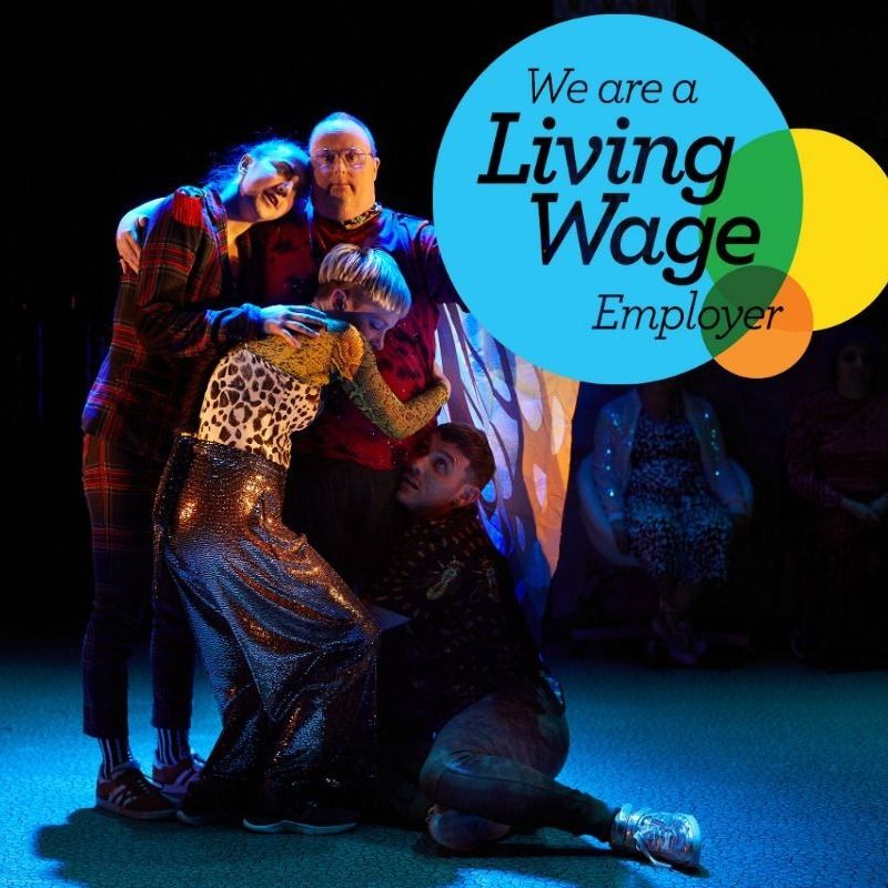 Production photography from Lung Ha show We Are All Just Little Creatures showing Ensemble member Keith Watson being embraced my dancers from Curious Seed theatre company. The Living Wage Employer mark is also displayed, featuring intersecting blue, yellow and orange circles and the text "We are a Living Wage Employer" © Peter Dibdin