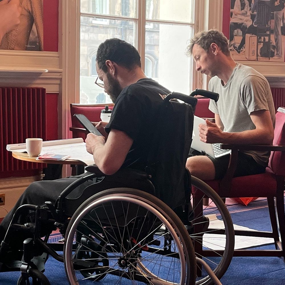 I'm Me Lead Artist Gavin and Lead Facilitator Ryan preparing for their appearance at the I'm Me Festival. They sit around a table going over notes written on flipchart paper, a phone and a laptop.