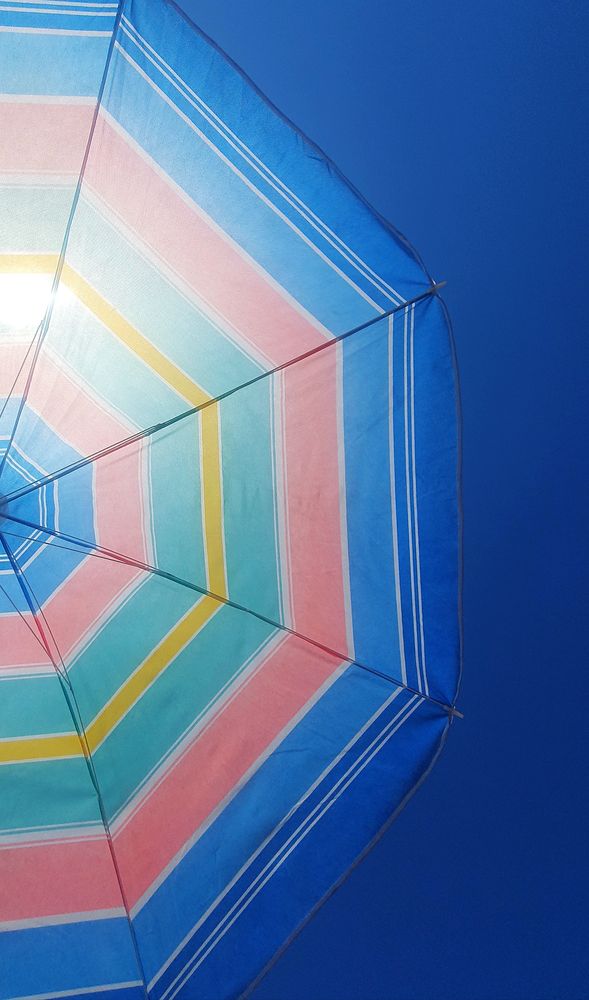 Ein rosa, blau, grün, gelb gestreifter Sonnenschirm. Perspektive von unten, man sieht die Hälfte des Schirms und blauen Himmel. 