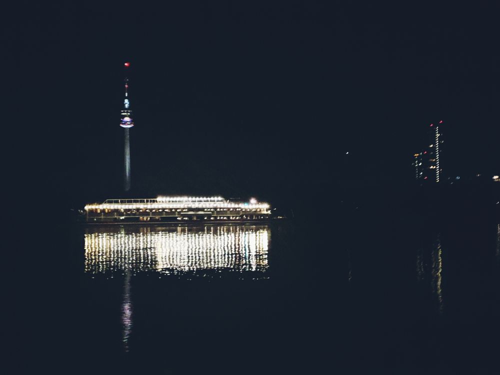Donau bei Nacht, ein Ausflugsschiff dessen Lichter sich im Wasser spiegeln ist zu sehen. Im Hintergrund ist außerdem der Donauturm zu erkennen. 
