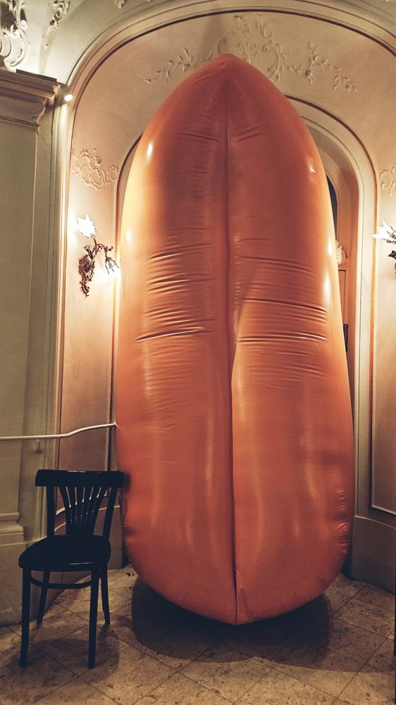 Im Foyer eines Theater steht eine etwa zwei Meter hohe aufblasbare Zunge, die seitlich von zwei Lampen beleuchtet wird. Links neben der Zunge steht ein schwarzer Sessel.