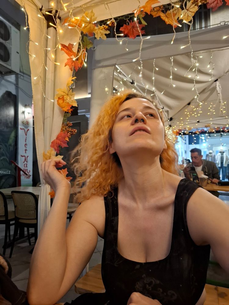 A woman with orange hair in a green corset sitting at an outside dining area with a background of lights and orange leaves.