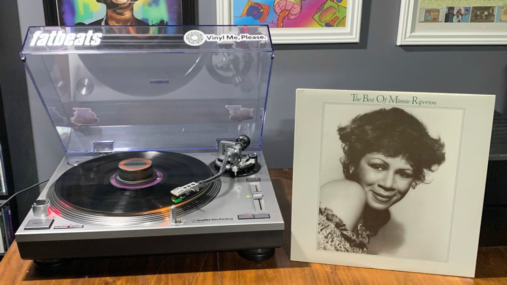 a vinyl pressing of “The Best of Minnie Riperton” playing on a turntable with the album cover (a black & white photo of Minnie with the album title printed in green just above it) propped up to the right