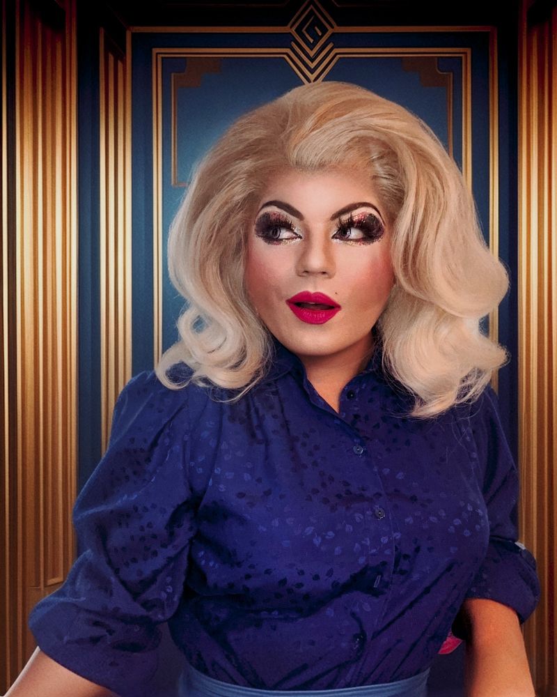 Eine blonde Drag Queen Anfang 40 mit üppiger Figur sitzt mit süffisant sinnlichem Blick vor einer blau goldenen Artdeco Wand. Sie trägt eine blaue aus Seidencrepe ihr Look erinnert an eine schicke Dame 