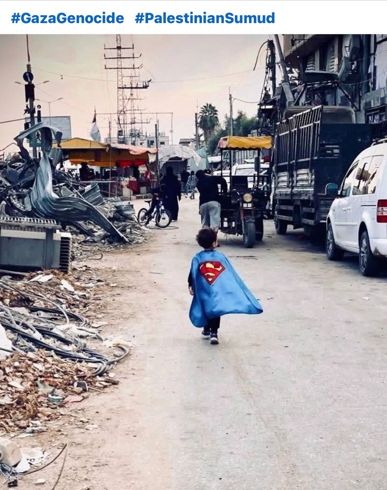 Little Palestinian child wearing a superman cape walks through the ruins of a genocide. 