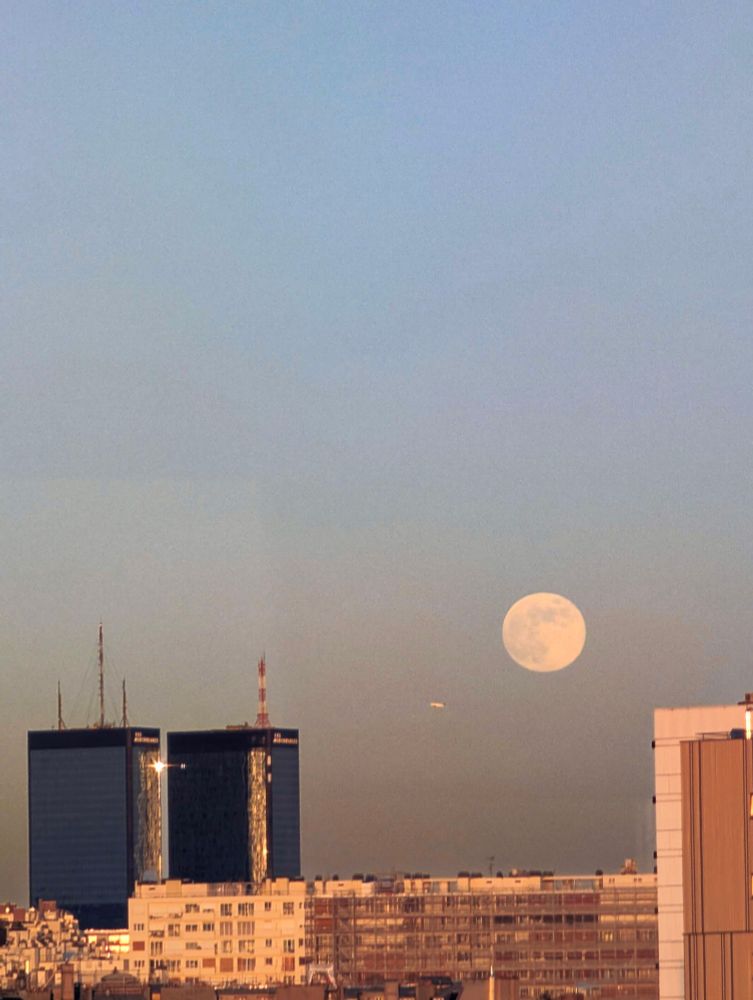 La pleine Lune se lève à côté des Mercuriales, qui reflètent encore le Soleil, alors qu'on devine un avion passe en dessous de la Lune