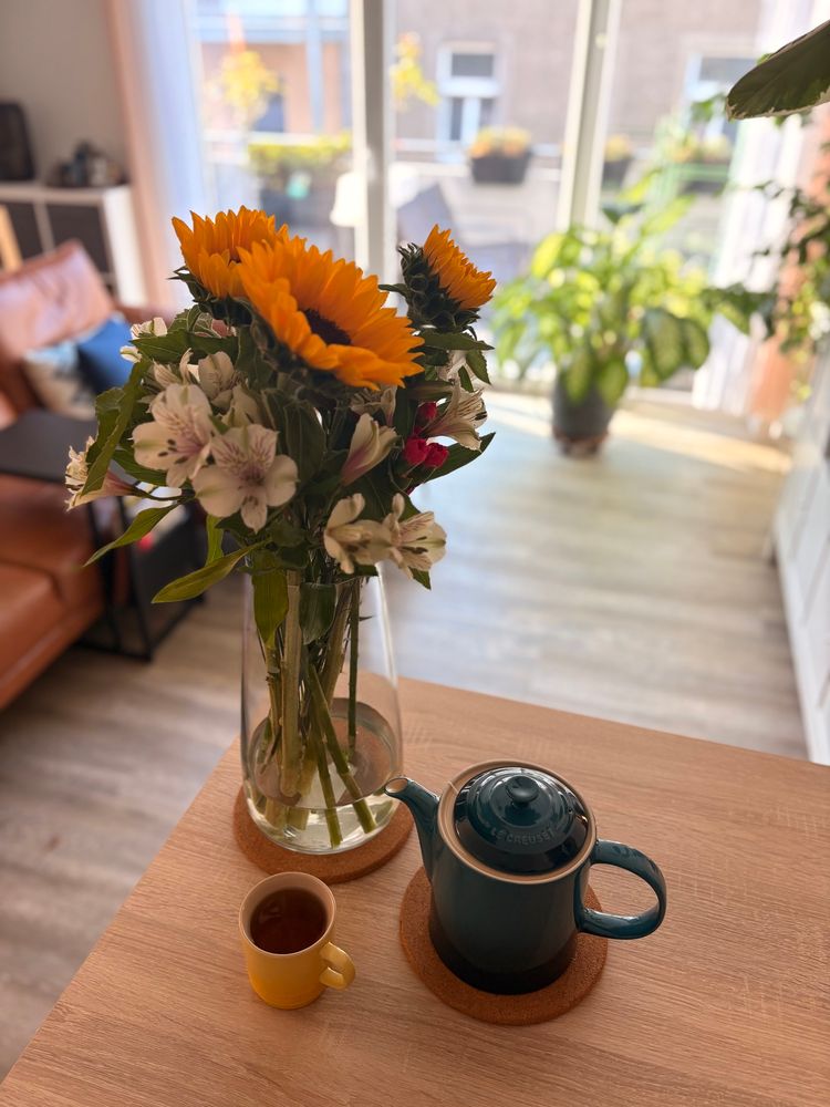 Ein Blumenstrauß mit Sonnenblumen und weißen und pinken Blüten auf einer Küchentheke, daneben eine blaue Teekanne und eine gelbe Teetasse. Im Hintergrund ein Wohnzimmer, in das die Abendsonne fällt. 