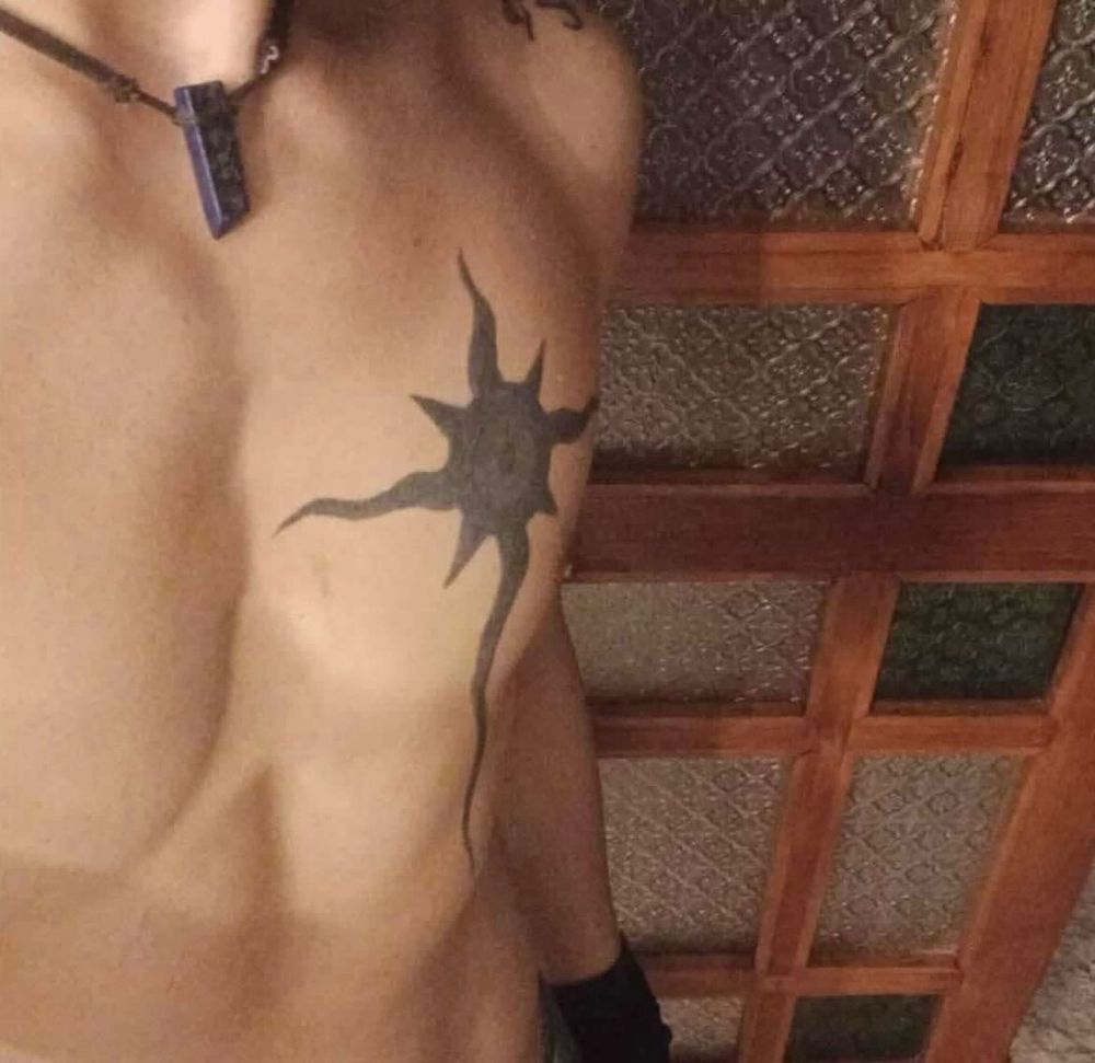 a person’s upper body before starting hormone replacement therapy (HRT), 11 months prior. They have a prominent tattoo of a black sun-like symbol on their chest and are wearing a necklace with a lapis lazuli stone pendant. The chest appears flat with visible muscle definition.