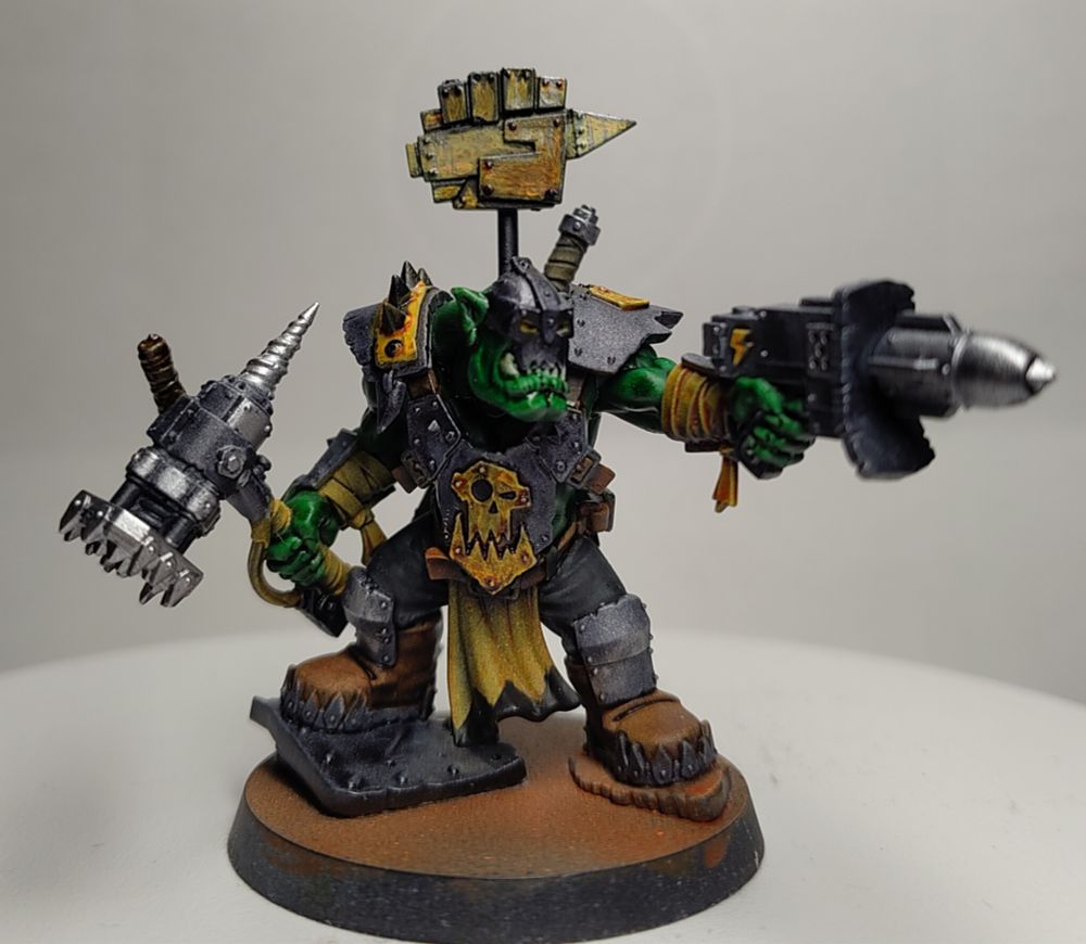 A Warhammer 40000 Space Ork miniature, a large Ork Nob with a spiked pneumatic hammer and a rocket launcher pistol, wearing metal armor painted yellow with black highlights in places