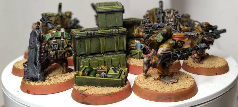 Turntable photo of a Warhammer 40000 Ratling Sniper Kill Team. Focus on the Stash master and a box of supplies