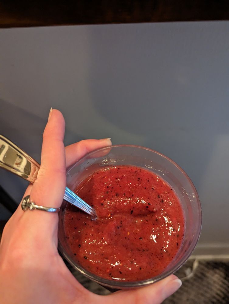 A glass of red smoothie with black flecks in it. The surface is textured as it is too thick to be liquid. There is a spoon in the glass.