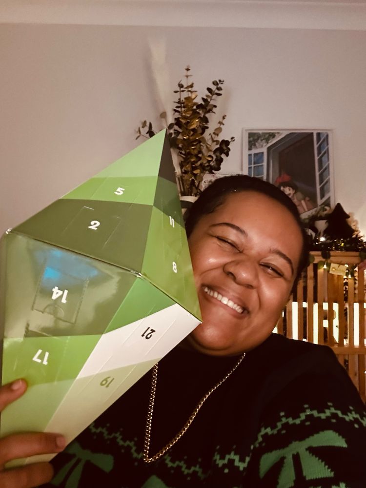 A picture of RiRi in a Christmas jumper holding a green plumbob shaped advent calendar
