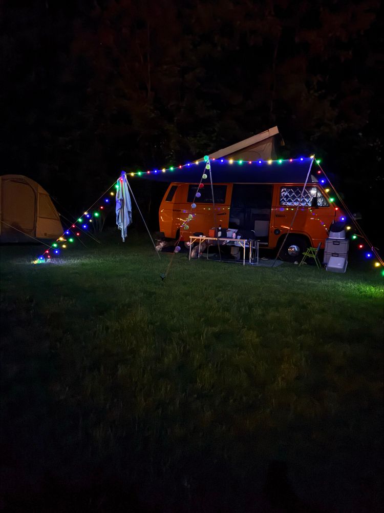 Campingbus voll aufgebaut auf Wiese in Nacht mit bunter Lichterkette.