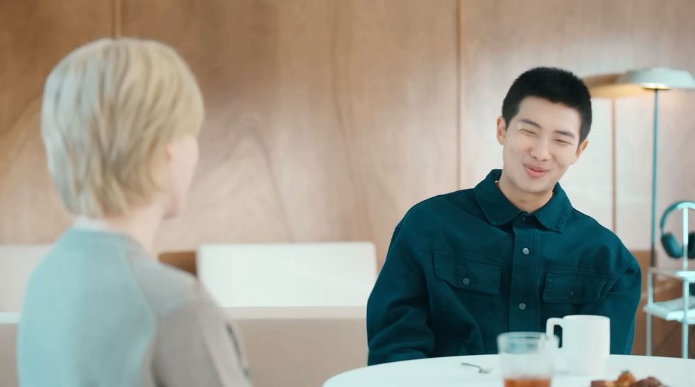 Screenshot from MMM( Mini&Moni Music) - Jimin video on BANGTANTV. Namjoon is in a Prussian blue/Indigo shirt, his hair is already trimmed for enlistment, and he is smiling. Jimin has his back towards the camera. He is in a beige sweater and his hair is dyed blond.