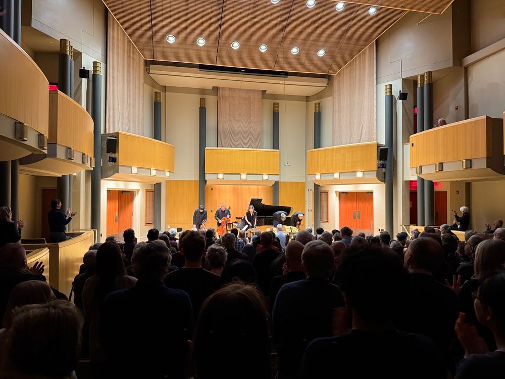 A recital hall with a full crowd applauding a string quartet and pianist onstage