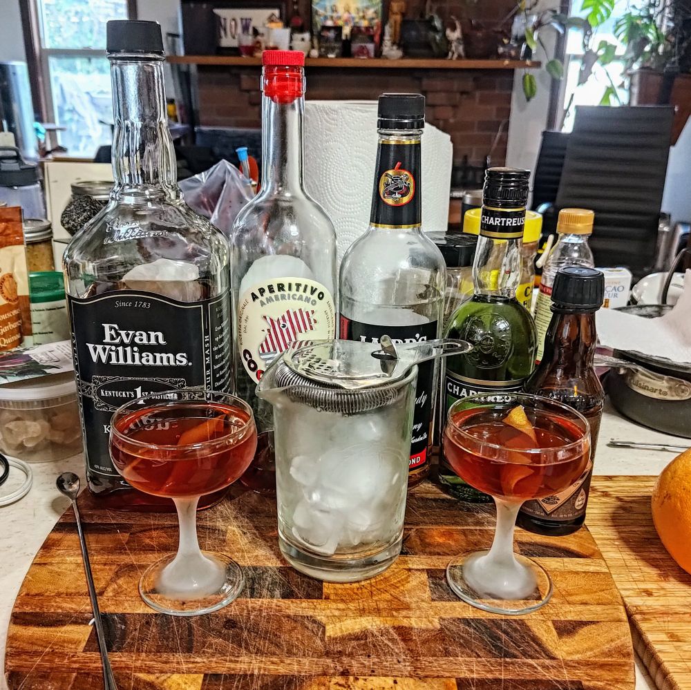 two reddish cocktails and a mixing glass in front of the bottled ingredients mentioned in the spec