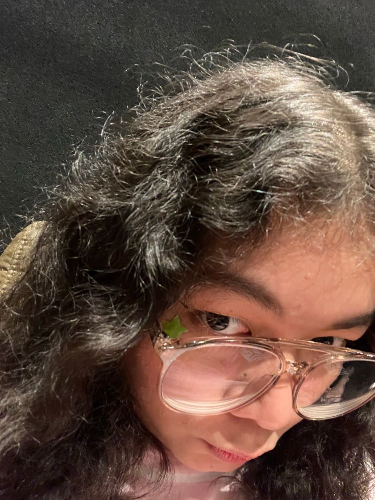 Up close selfie of a curly-haired woman with transparent framed glasses. She has a green star-shaped pimple patch on the corner her left eye.