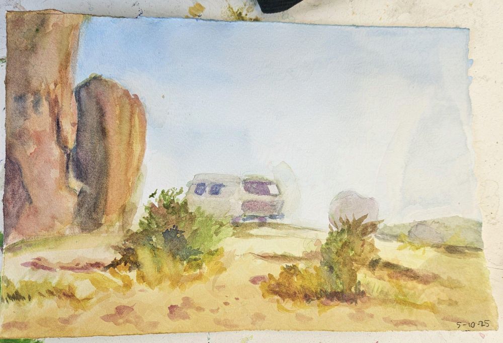 A watercolor landscape painting. It depicts a desert. Large rocks line the left of the page. Sand with shrubs are in the foreground. In the far background is a van