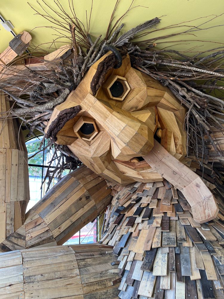 A giant wooden troll is seated on the floor. Her head is tilted and still touches the ceiling. A huge tongue extends from her mouth and down her belly