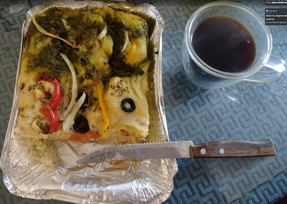 Picture of a Focaccia, it has olives, peppers, onion, small tomatoes and pesto as toppings. There is also a small cup of coffee on the side