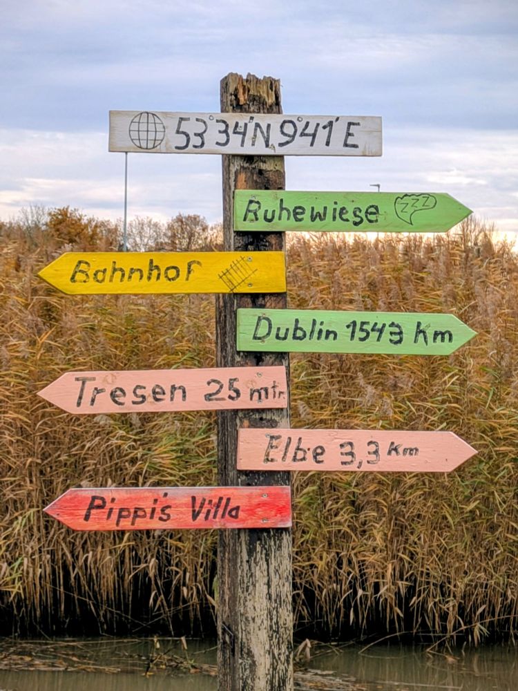 A wooden signpost pointing at various wildly different locations, including "Dublin 1543km", "Elbe 3.3km" and "Tresen 25m" 