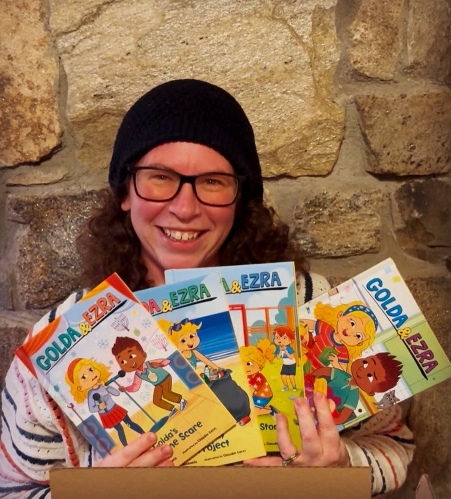 A photo of me - Hollie wearing a black hat, smiling ear to ear and holding four Golda and Ezra books - chapter book series. 