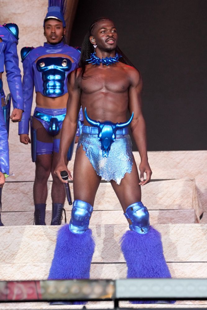 lil nas x, a Black artist and singer, performing topless with blue tinsel around his neck, a blue glittery short and asymmetrical skirt, and a belt with a goat head with large horns on it. he also has blue knee pads and fur across his shins likely on his shoes. behind him is a Black back-up dancer with a blue hat with fringing at the top, a long sleeve blue top with arched shoulder pads and a shiny blue six back and pecks/breasts with nipples, and shorts which are cut out like underwear with a shiny penis on them and short dark blue boots. the stage is a cream or beige stone colour. 