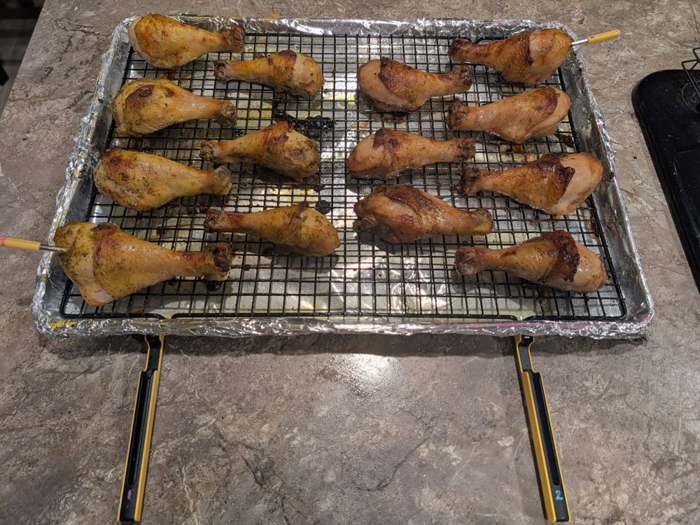 A foil-lined sheetpan with a shmultz-laden wire rack holding chicken drumsticks with temperature probes for 2 of the largest drumsticks in each half. The left half is seasoned with lemon pepper while the other is seasoned with buttermilk ranch. Both sides have internal temps held at 155⁰ for 30 min sections of crispy skin for textural variety.