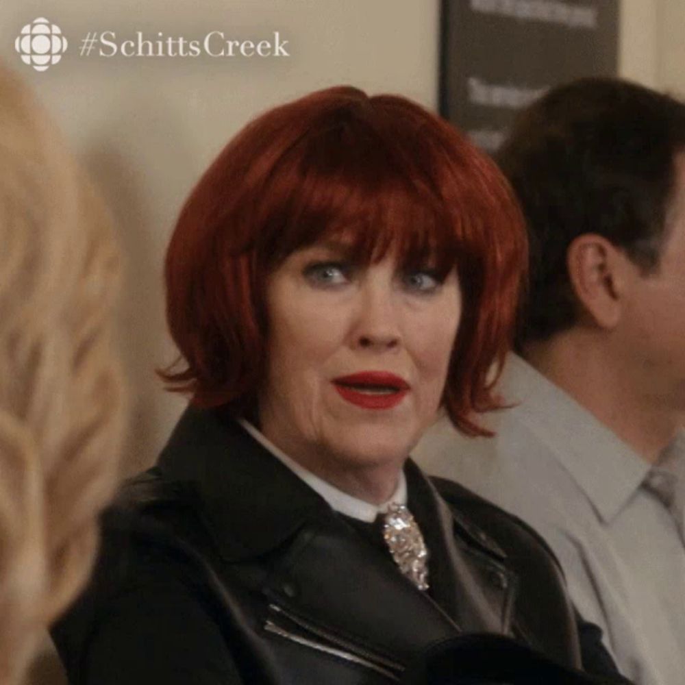 a woman with red hair is wearing a black jacket and a #schittscreek logo behind her