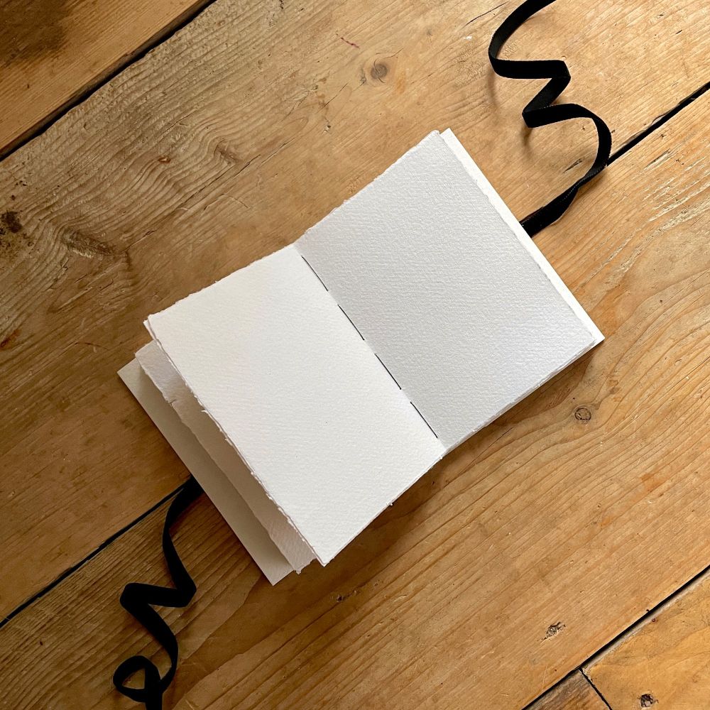 Open (blank) pages of Cotton Rag Sketchbook stitched in black thread lies on natural wood surface; a black velvet ribbon curls out from either side of the book.