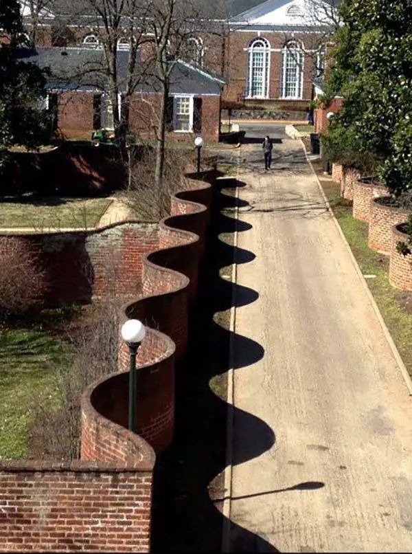 I think this is University of Virginia's gardens near the main forum building, showing a red brick wall that is serpentine in shape, casting an odd undulating shadow on the white-paved road/driveway nearby.