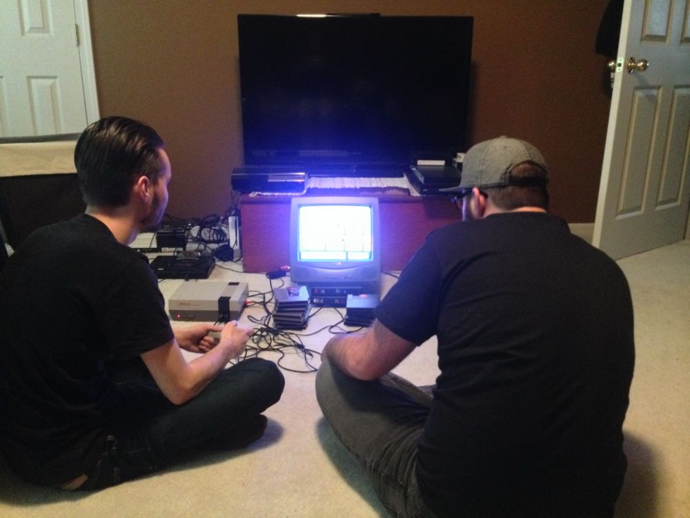 Ryan and Patrick sitting cross legged in a bedroom in front of a small CRTV playing Nintendo Entertainment System. A blown-out screen is on the TV.