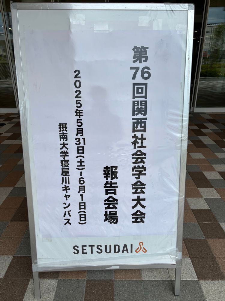 摂南大学寝屋川キャンパスの建物入口前に設置された、第76回関西社会学会大会の報告会場を示す案内看板。開催日時は2025年5月31日（土）〜6月1日（日）。

A signboard in front of a building entrance at Setsunan University Neyagawa Campus indicating the venue for the 76th Kansai Sociological Society Conference. The event is scheduled for May 31 (Saturday) to June 1 (Sunday), 2025.