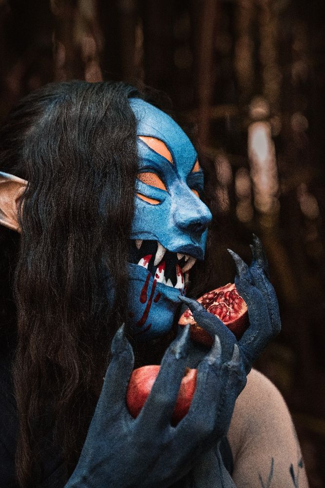 close up portrait of my blue time war cosplay, in a blue mask with 6 yellow eyes, holding  two halves of a pomegranate up to my mouth. There is blood on the teeth and chin of the mask.