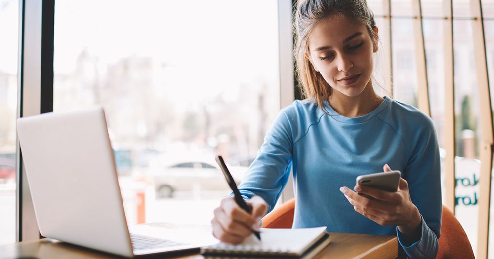 Eine junge Frau sitzt vor ihrem Laptop und schaut auf ihr Smartphone.