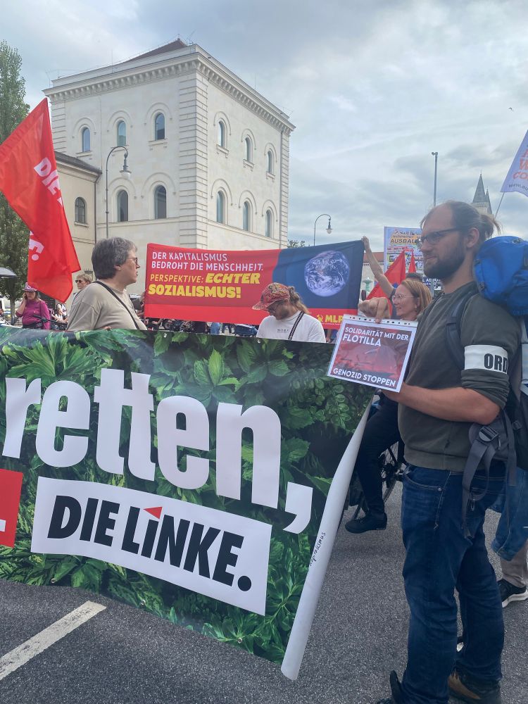 Während eine Demo in München, Menschen mit Transpi der Linkspartei halten auch ein A4-Blatt, wo drauf steht Solidarität mit der Flotilla - Genozid stoppen. Eine Transpihalterin ist Brigitte Wolf, Stadträtin in München 