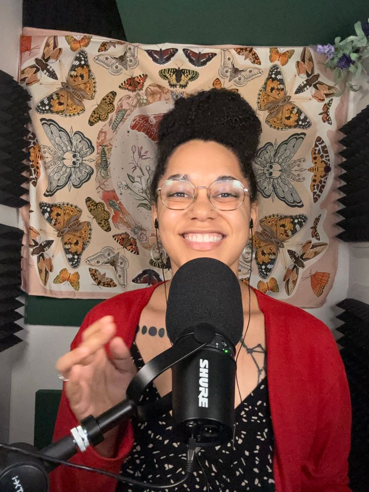 Moiya smiling behind a microphone in her recording closet with sound dampening boards on the walls and a moth-themed cloth hanging behind her 