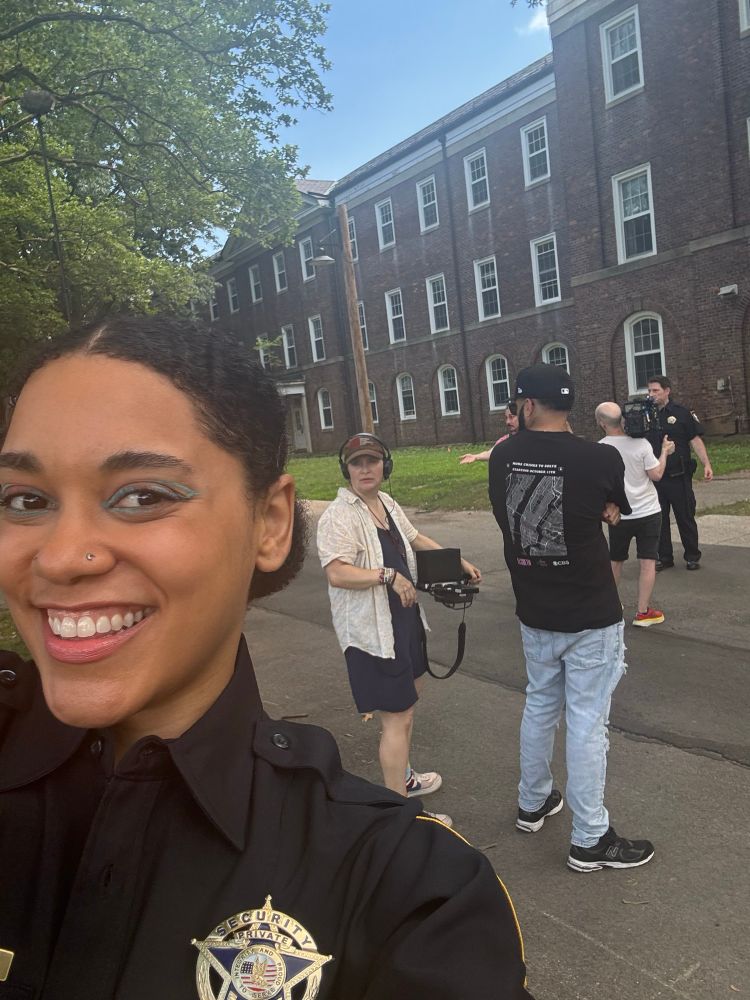 Selfie of Moiya smiling in a security guard costume. Behind her there’s a small camera crew filming another person in a security guard costume, all standing on a paved road in front of a long brick building with many windows 