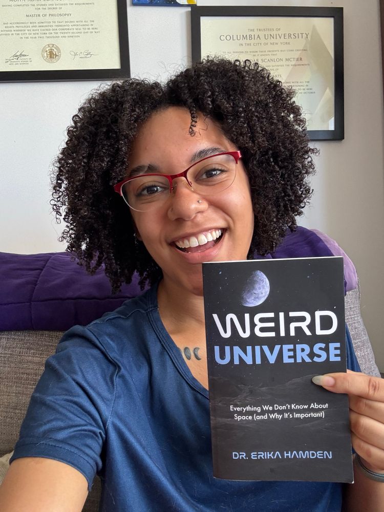 Selfie of Moiya smiling and holding up a copy of Weird Universe by Dr. Erika Hamden 