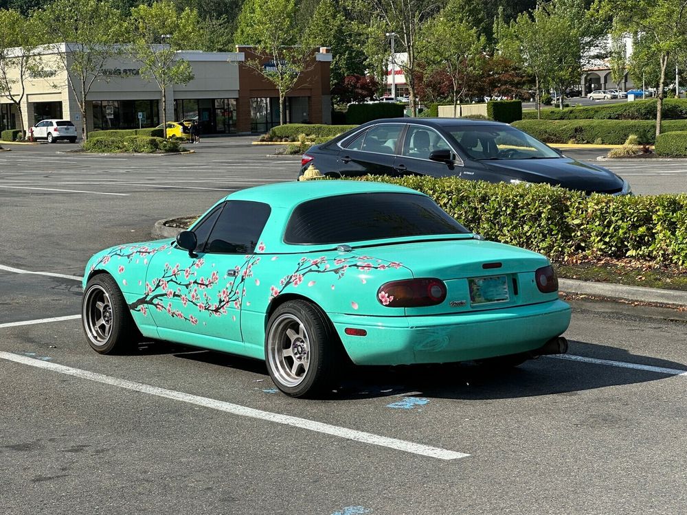 a miata parked in a parking lot with a greenish-teal paintjob and cherry blossoms painted on in the style of Arizona Iced Tea with Ginseng and Honey's packaging