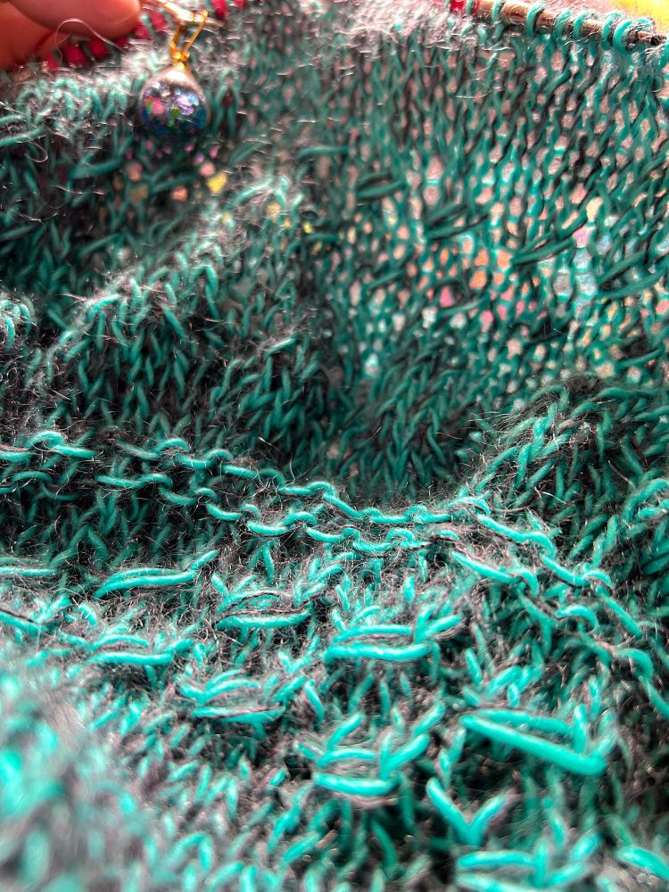A close up of the jumper being knitted in hand dyed yarn held double, showing some cosy fuzzy halo - one strand of aqua blue fingering weight Merino/Mohair brushed singles, and one strand of charcoal grey lace Mohair/Silk, creating a mottled effect. There’s a mix of lace patterns and stockinette in the knitted fabric.