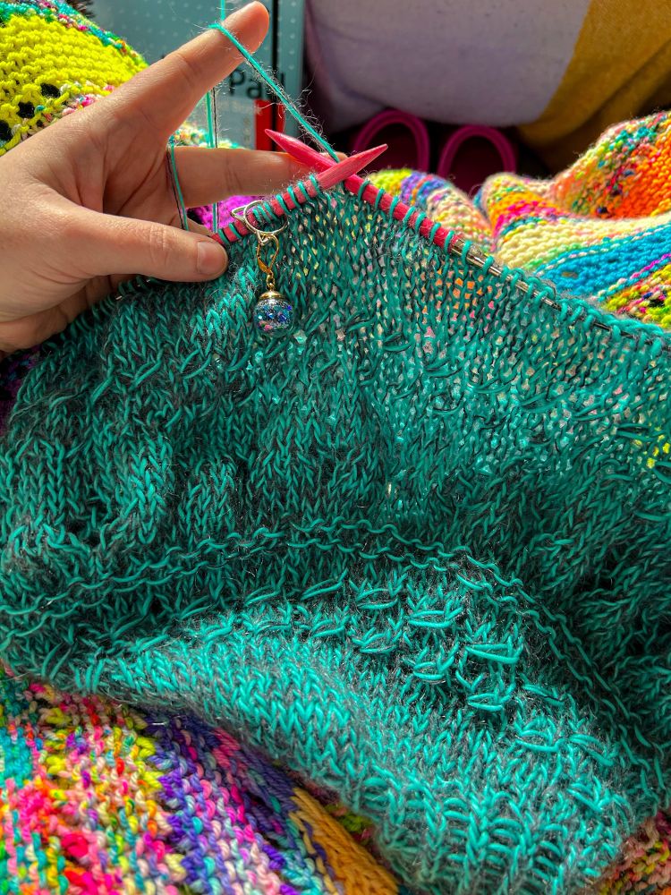 Tracey is holding a jumper WIP on circular needles in her left hand, index finger held out continental style. The needles are pink, and there’s a bauble stitch marker filled with blue glitter hanging off a cat head shaped ring on the left needle. The jumper is being knitted in hand dyed yarn held double - one strand of aqua blue fingering weight Merino/Mohair brushed singles, and one strand of charcoal grey lace Mohair/Silk, creating a mottled effect (is mottled the right word? I dunno but it’ll suffice! 🥰). There’s a mix of lace pattern and stockinette in the knitted fabric. Underneath the WIP is Tracey’s recently finished Habitation Throw, which is knitted in brightly coloured stripes using scraps of DK.