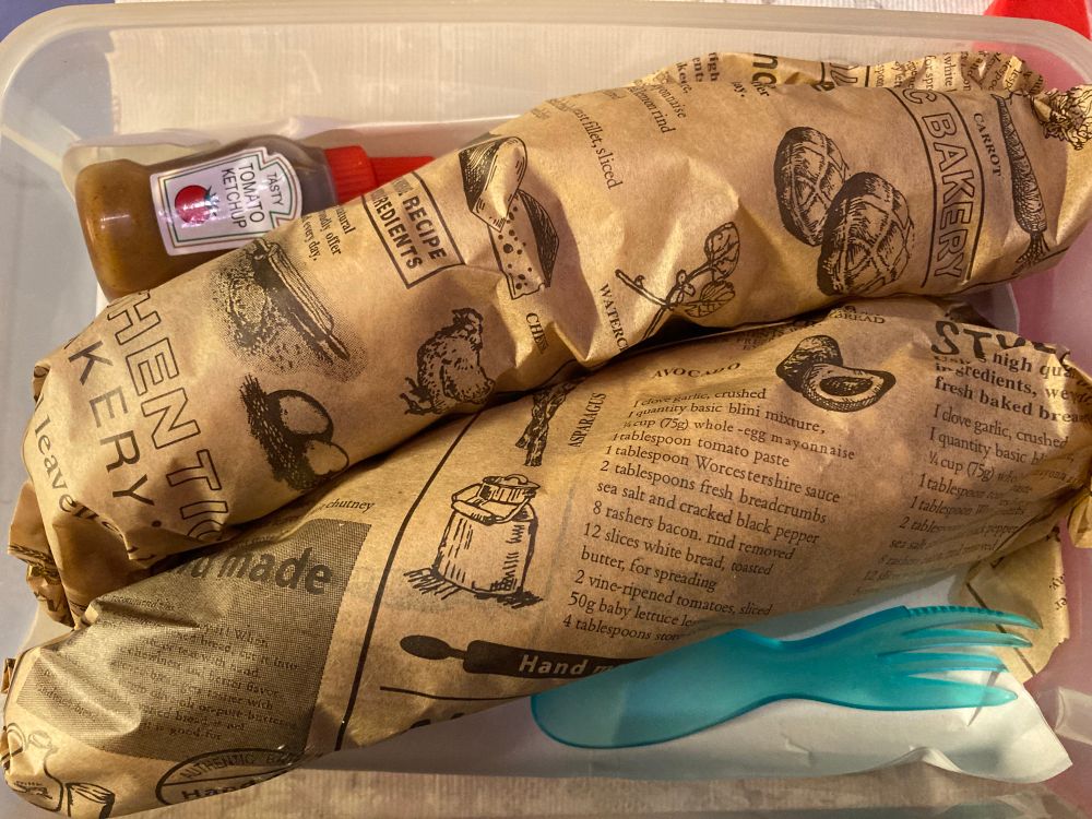 Beide in bedrucktes Delipapier gewickelte Käselaugenstangen dazu eine kleine Quetschflasche mit Gewürzketchup und ein Göffel